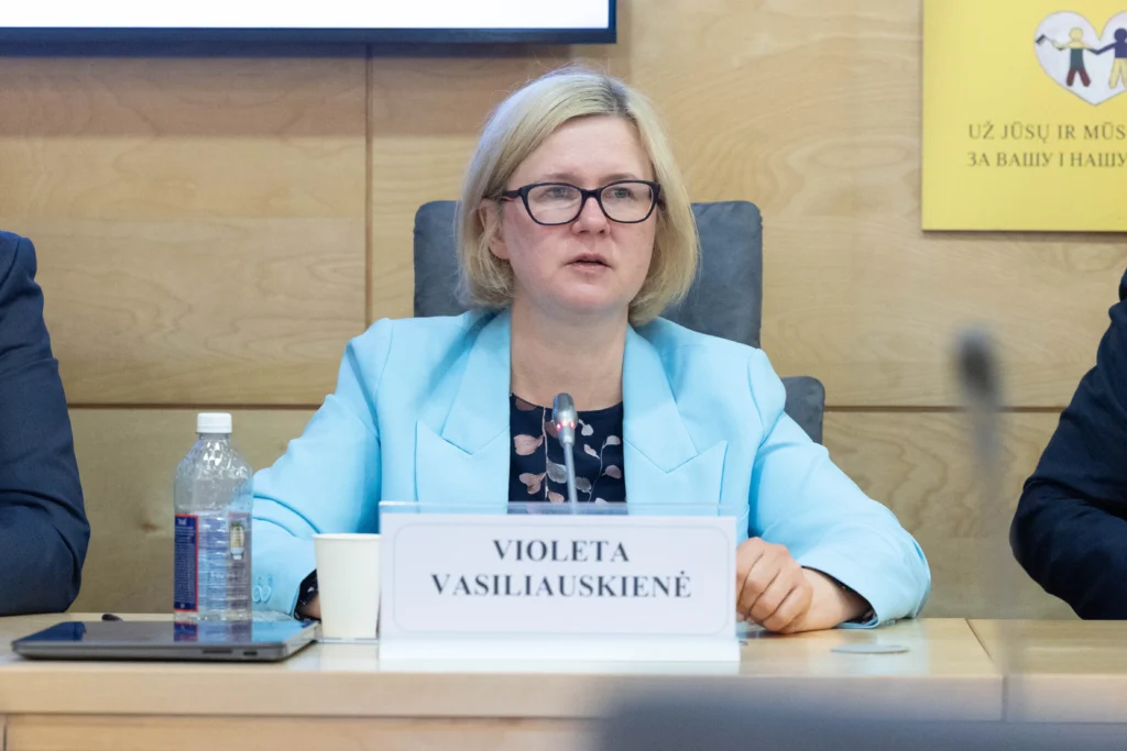 Trumpaplaukė moteris su akiniais ir žydru švarku sėdi prie stalo su savo vardine kortele.