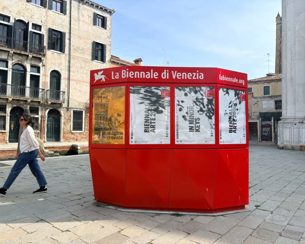 Raudonas „La Biennale di Venezia“ informacinis stendas su parodų plakatais aikštėje, pro kurį praeina moteris.