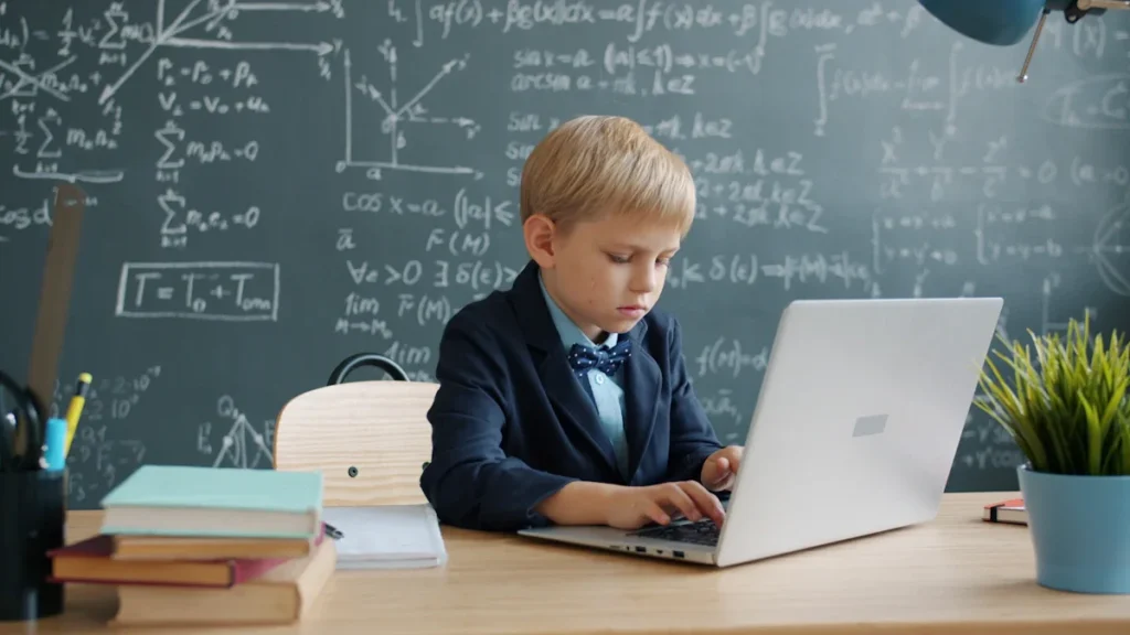 Mokinys prie stalo su kompiuteriu ir lenta su formulėmis klasėje.