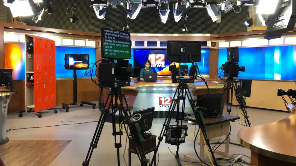 Televizijos žinių studija su profesionaliomis kameromis ir apšvietimo įranga, reprezentuojanti transliuotojo darbo aplinką.