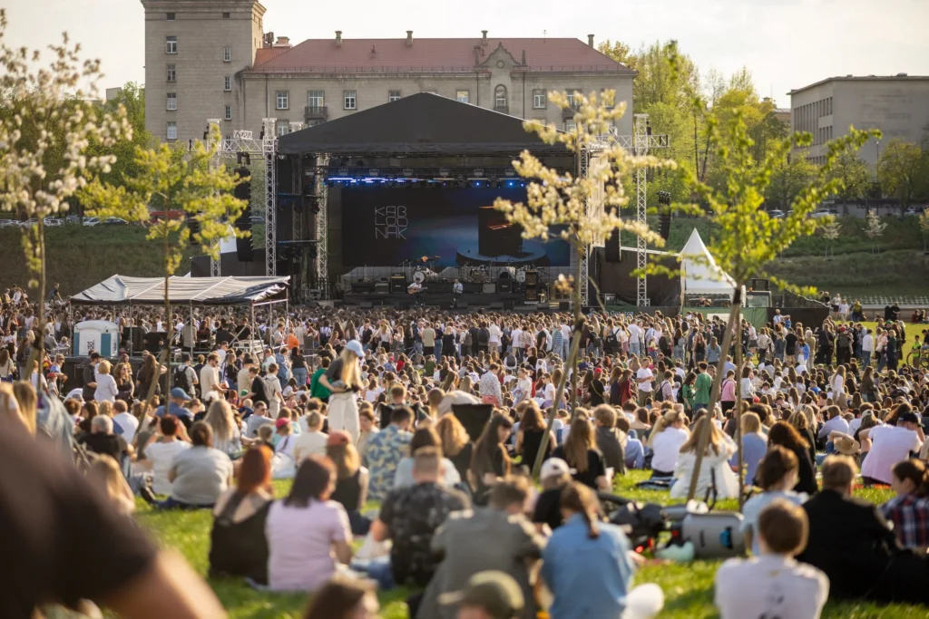 Didelė žmonių minia sėdi ant žolės priešais didelę koncertinę sceną saulėtą popietę parke.