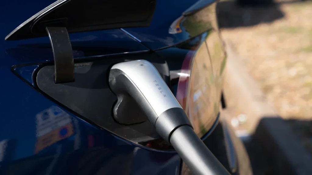 Mėlynos spalvos elektromobilis įkraunamas viešoje stotelėje, naudojant standartinę įkrovimo jungtį saulėtą dieną.