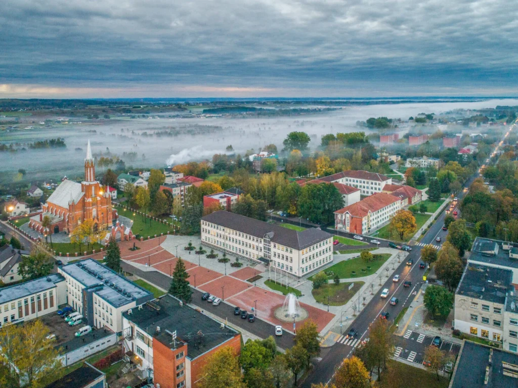 Iš paukščio skrydžio matoma raudonų plytų bažnyčia, miesto aikštė su fontanu ir tirštas rūkas fone.