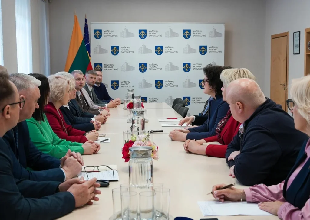 Grupė žmonių sėdi prie ilgo stalo Mažeikių rajono savivaldybės posėdžių salėje susitikimo metu.