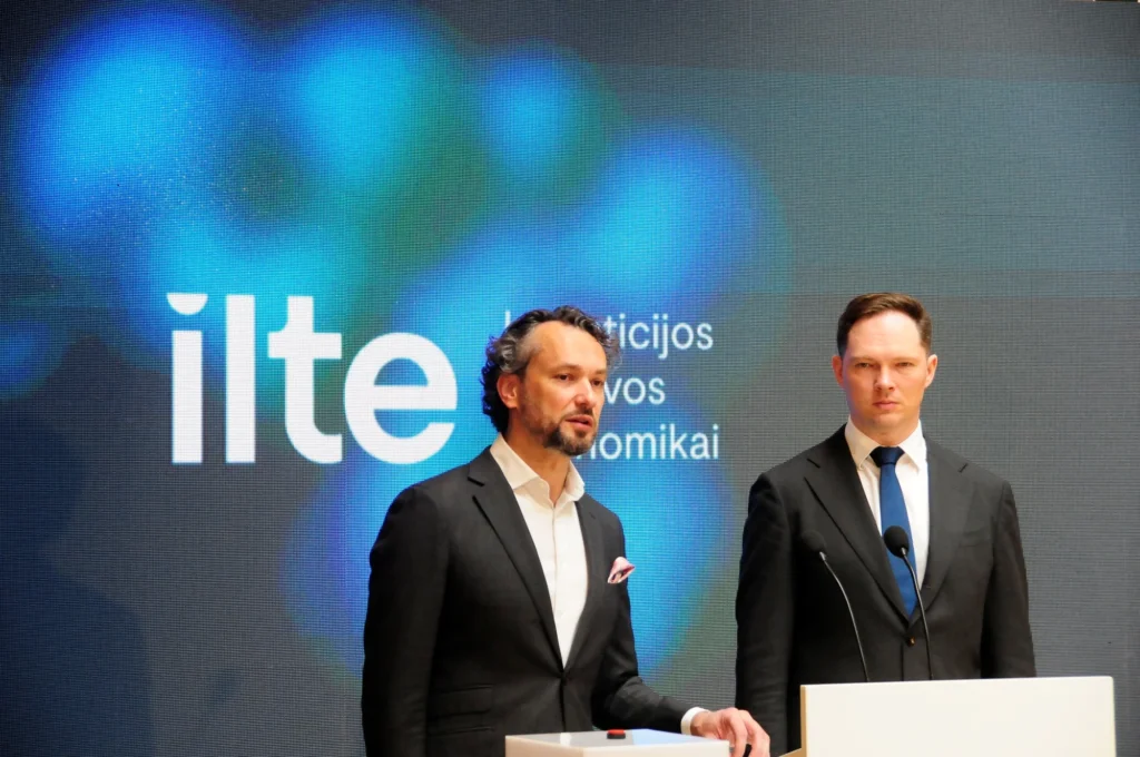 Du vyrai tamsiais kostiumais stovi priešais didelį ekraną su užrašu „ilte“ ir sako kalbą.