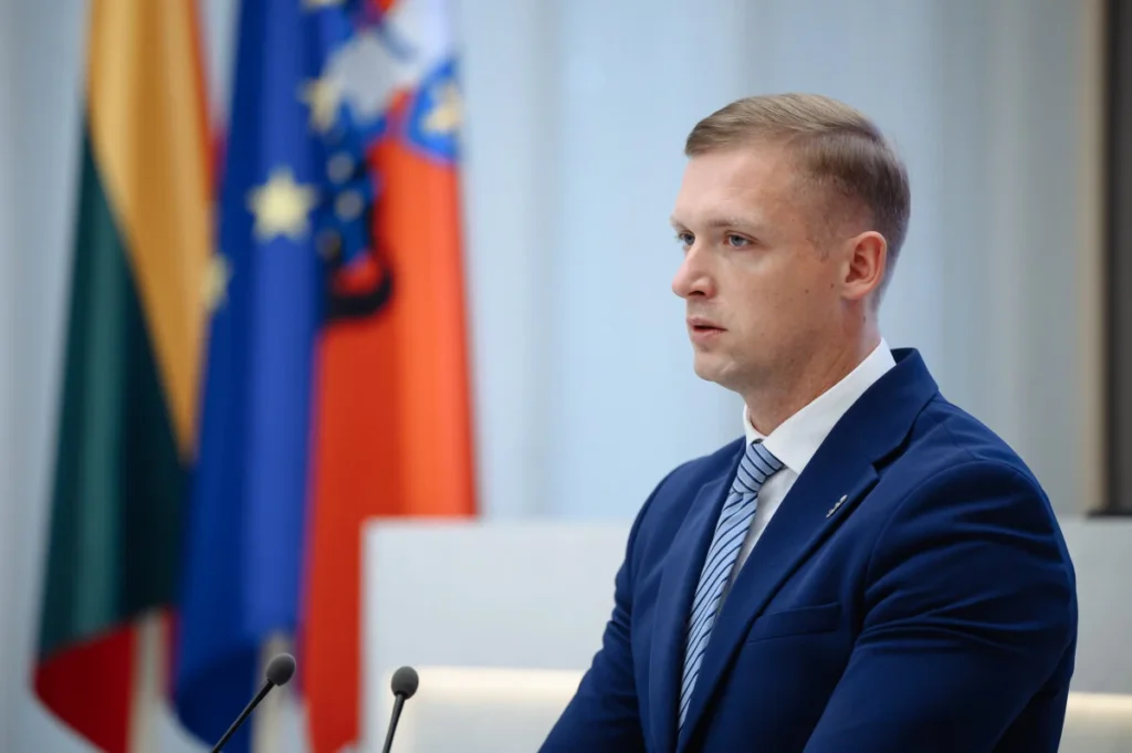 Mindaugas Lingė su mėlynu kostiumu kalba prie mikrofono Lietuvos ir Europos Sąjungos vėliavų fone.