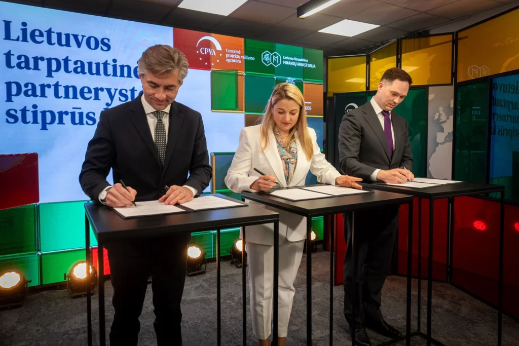 Trys asmenys dalykiniais kostiumais pasirašo dokumentus fone esant ekranui su Lietuvos valstybinių institucijų logotipais.