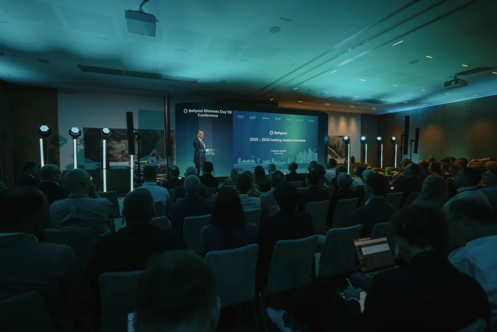 Pranešėjas prie pulto skaito pranešimą sėdinčiai auditorijai konferencijų salėje su dideliu ekranu.