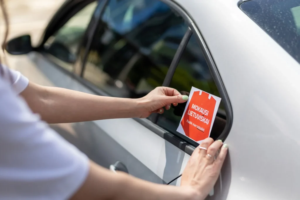 Rankos klijuoja raudoną lipduką su užrašu „Mokausi lietuviškai“ ant pilko automobilio galinio lango.