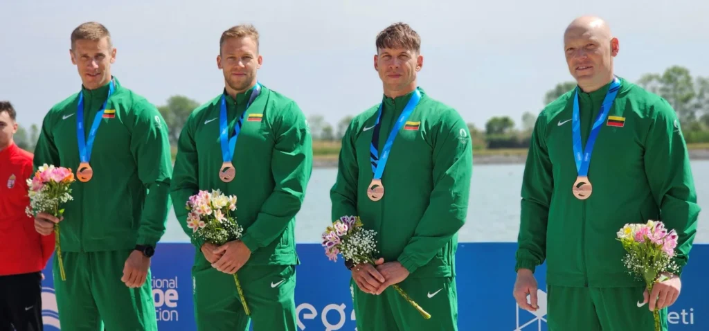 Keturi sportininkai su žaliomis Lietuvos rinktinės aprangomis, pasipuošę bronzos medaliais ir laikantys gėlių puokštes.