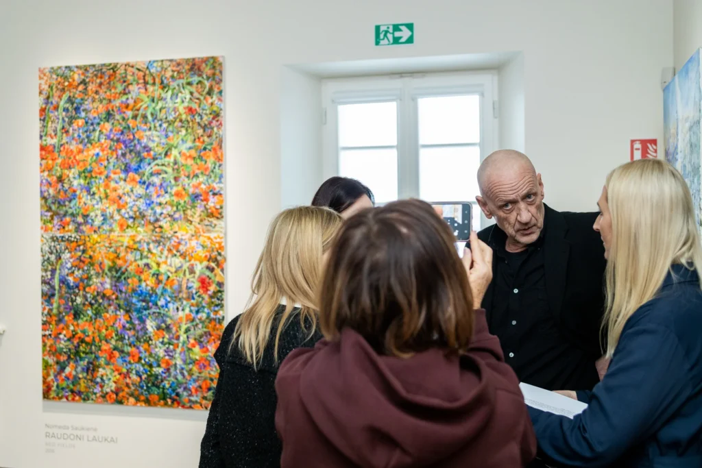 Meno galerijoje lankytojai stebi ryškų paveikslą, o viena moteris jį fotografuoja telefonu.