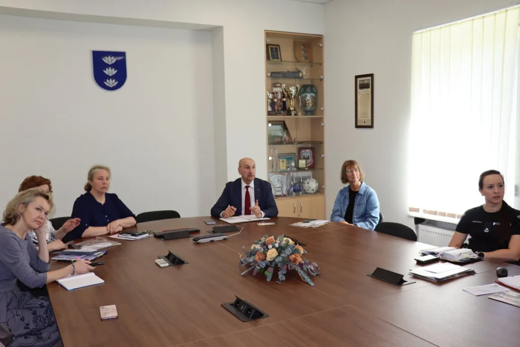 Grupė žmonių sėdi prie didelio ovalaus stalo dalykiškame posėdyje šviesiame kabinete su vėliava.