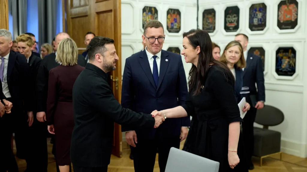 Ukrainos prezidentas Volodymyras Zelenskis spaudžia ranką Viktorijai Čmilytei-Nielsen formalioje aplinkoje tarp kitų pareigūnų.