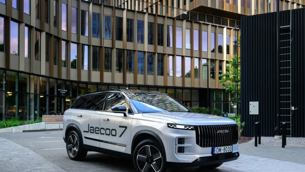 Baltas „Jaecoo 7“ visureigis stovi šiuolaikinio pastato fone, su matomu latvišku automobilio numeriu.