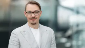 Šviesiaplaukis vyras su akiniais ir šviesiu švarku pozuoja šviesiame, neryškiame biuro fone.