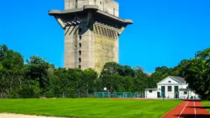 Didelis betoninis bokštas virš sporto aikštelės su bėgimo takais bei žalia veja.