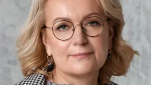 Vidutinio amžiaus šviesiaplaukė moteris su akiniais ir languotu švarku pozuoja pilkame fone.