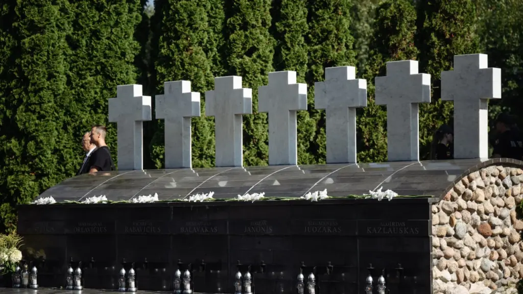 Eilė baltų kryžių virš tamsaus paminklo su išgraviruotais vardais ir padėtomis baltomis gėlėmis.