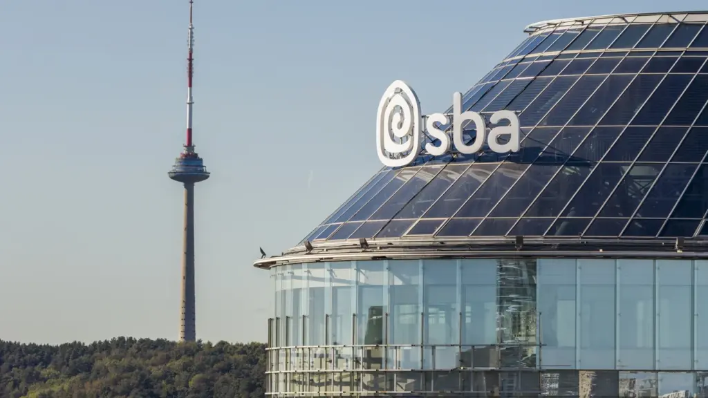 Modernas stiklinis pastatas su „sba“ logotipu ir televizijos bokštas Vilniaus panoramoje.