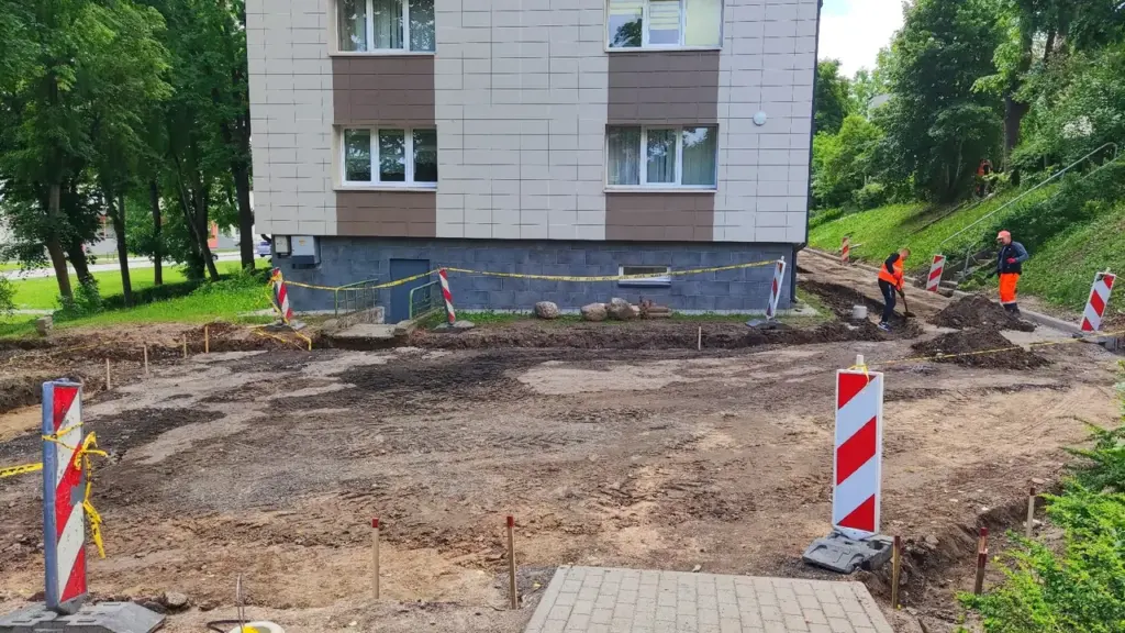 Du darbininkai su ryškiomis liemenėmis tvarko žemę šalia daugiabučio namo, aptverto įspėjamąja juosta.