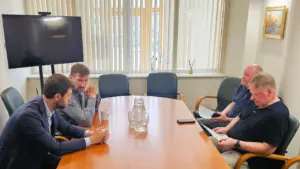 Keturi vyrai sėdi aplink ovalų medinį stalą biuro susitikime šviesiame kabinete su žaliuzi.