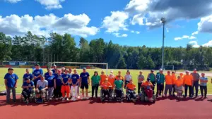 Grupė žmonių ir neįgaliųjų vežimėliuose sporto stadione saulėtą dieną po atviru dangumi.