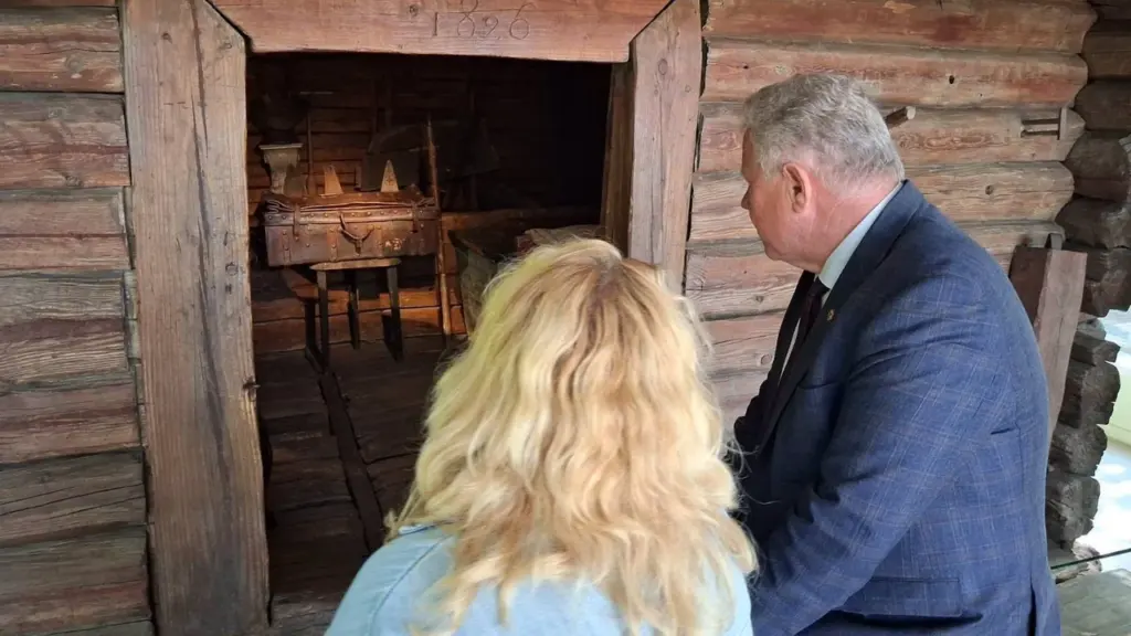 Vyras su kostiumu ir moteris žiūri į senovinį rąstinį namą su 1826 metų įrašu.