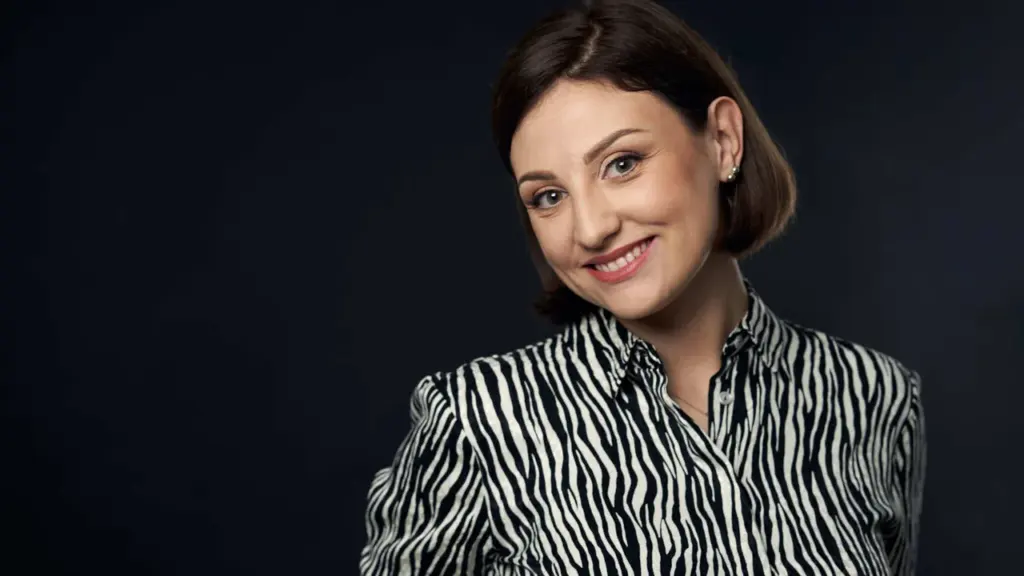 Besišypsanti tamsiaplaukė moteris su zebro rašto marškiniais tamsiame studijos fone, žiūrinti į kamerą.