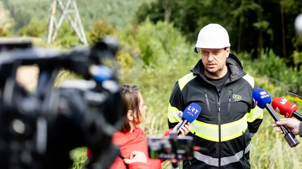 ESO darbuotojas su baltu šalmu duoda interviu žurnalistams lauke prie elektros linijos atramos.