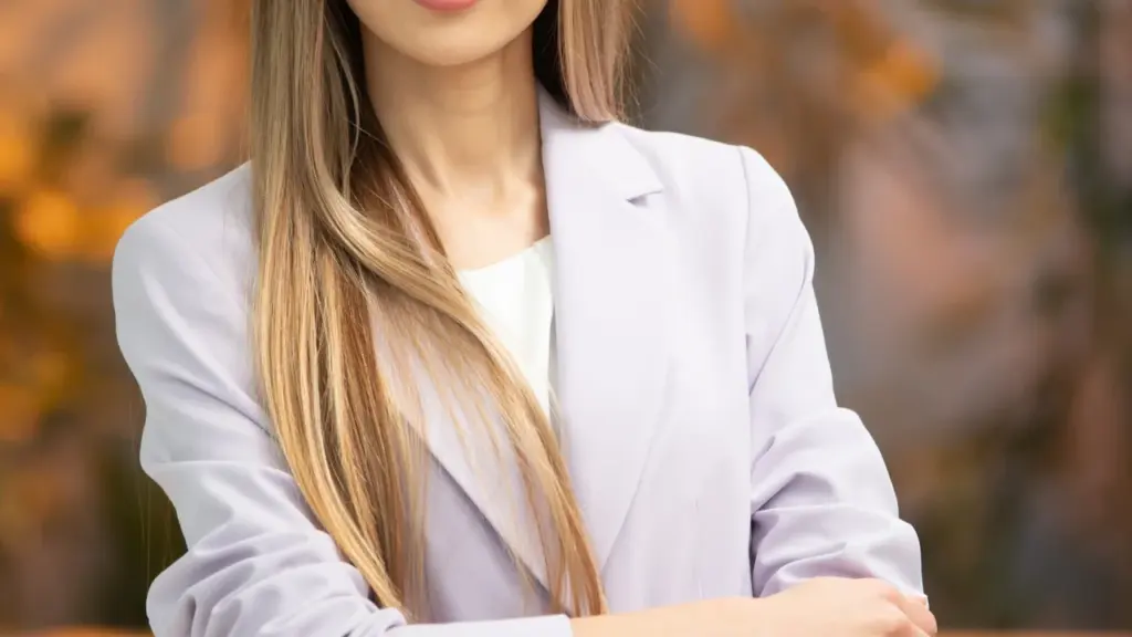 Šviesiaplaukė moteris šviesiai violetiniame kostiumėlyje stovi susikryžiavusi rankas ir žvelgia į kamerą.