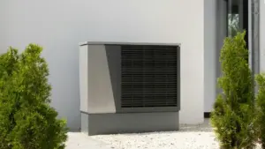Modernus pilkas šilumos siurblys stovi lauke prie baltos pastato sienos tarp tujų.
