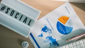 Darbalaukis su užrašų knygute, kompiuterio klaviatūra ir statistikos diagramomis apie socialinę pagalbą
