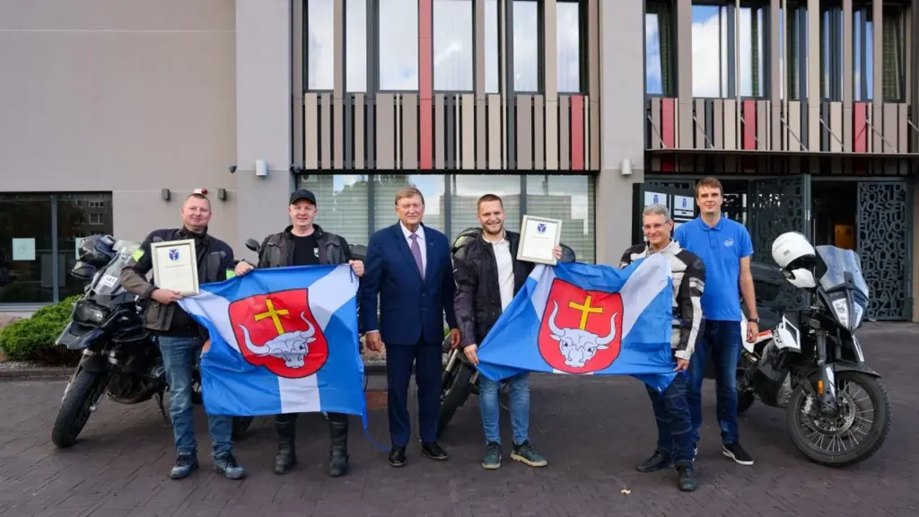 Grupė vyrų su motociklais laiko Kauno rajono vėliavą ir padėkos raštus prie modernaus pastato.