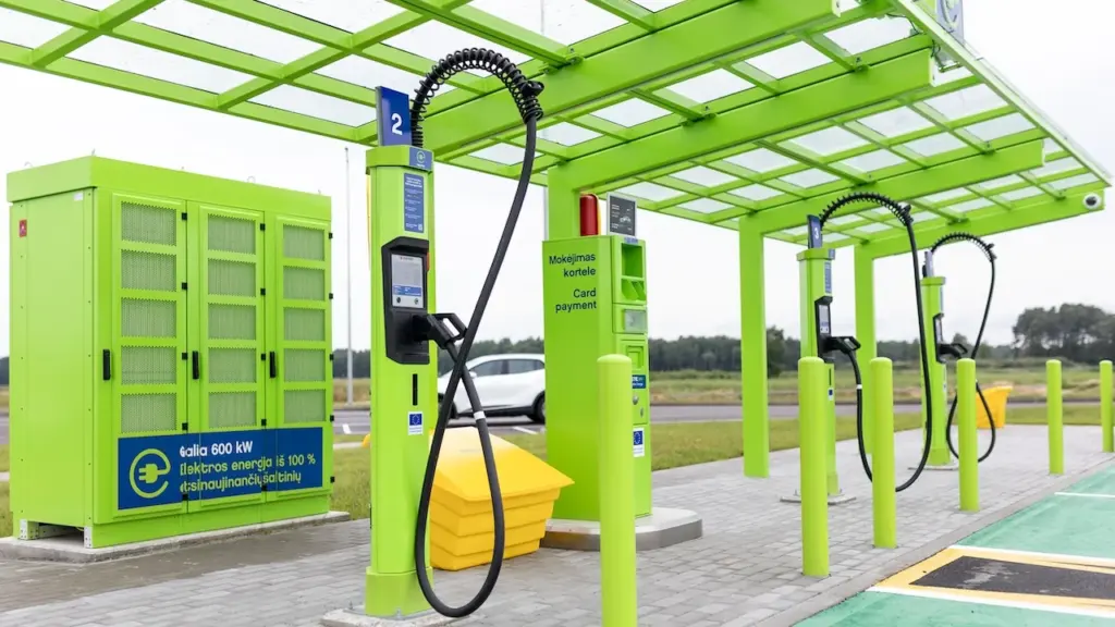 Šviesiai žalios spalvos elektromobilių įkrovimo stotelės su laidais ir stogine atviroje lauko aikštelėje.