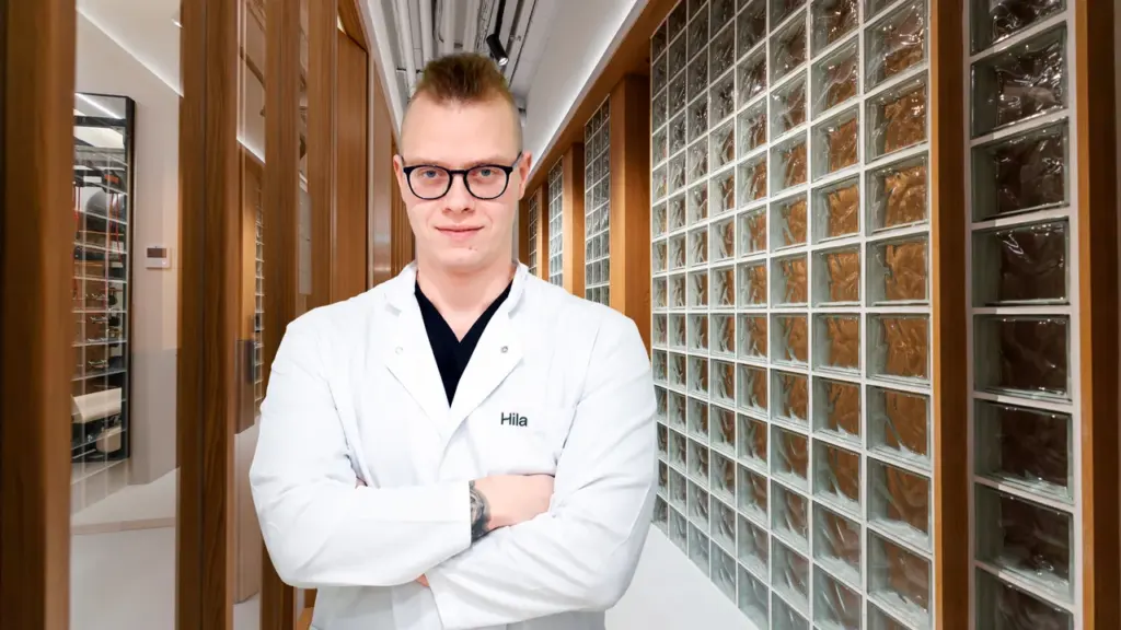 Jaunas vyras gydytojas su akiniais ir baltu chalatu stovi koridoriuje sukryžiavęs rankas.