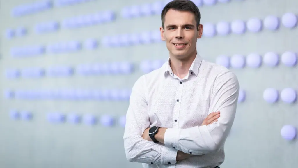 Besišypsantis vyras baltais marškiniais sukryžiuotomis rankomis stovi fone su šviečiančiais apvaliais elementais.