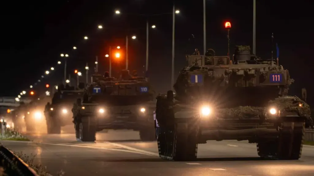 Kariniai tankai su įjungtais žibintais naktį važiuoja asfaltuotu keliu per tamsią gyvenvietę.