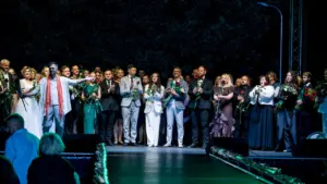 Grupė žmonių vakariniuose drabužiuose stovi scenoje, daugelis laiko raudonas rožes ir ploja renginio pabaigoje.