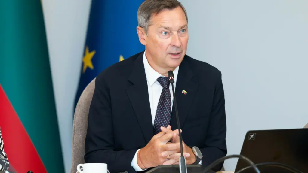 Vidutinio amžiaus vyras su kostiumu sėdi prie stalo priešais mikrofoną oficialiame renginyje.