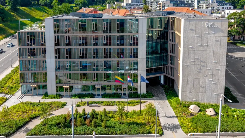 Modernas stiklinis verslo centras su žaliuojančiu sodu ir vėliavomis prie pagrindinio įėjimo