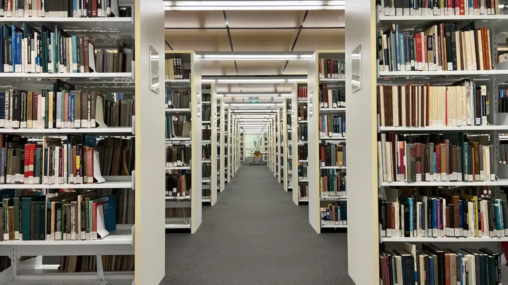 Tvarkingos bibliotekos lentynos su knygomis šviesioje ir erdvioje skaitymo erdvėje.