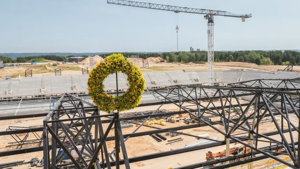 Didelis saulėgrąžų vainikas pritvirtintas prie statomo stadiono metalinės konstrukcijos, matoma statybvietė ir kranas.