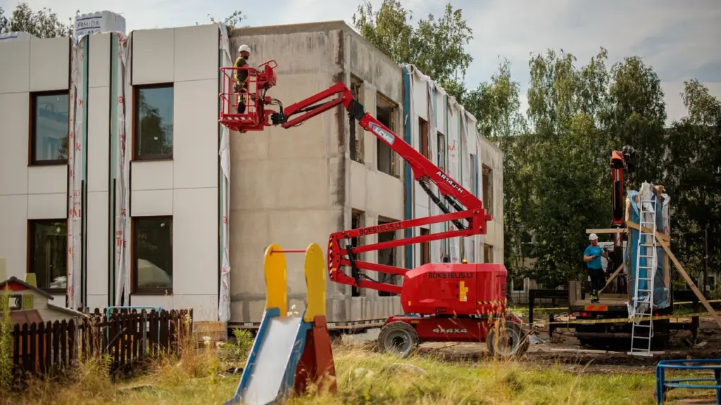 Statybininkas raudoname kėlimo mechanizme dirba prie renovuojamo pastato sienos šalia žaidimų aikštelės.