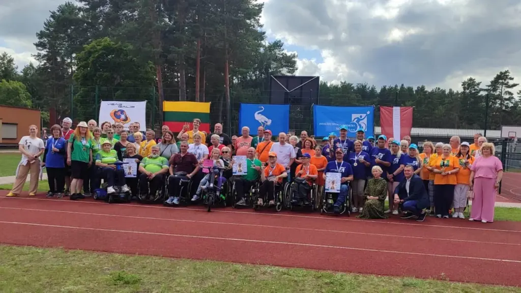 Didelė žmonių grupė su neįgaliojo vežimėliais pozuoja stadiono bėgimo takelyje su vėliavomis fone.