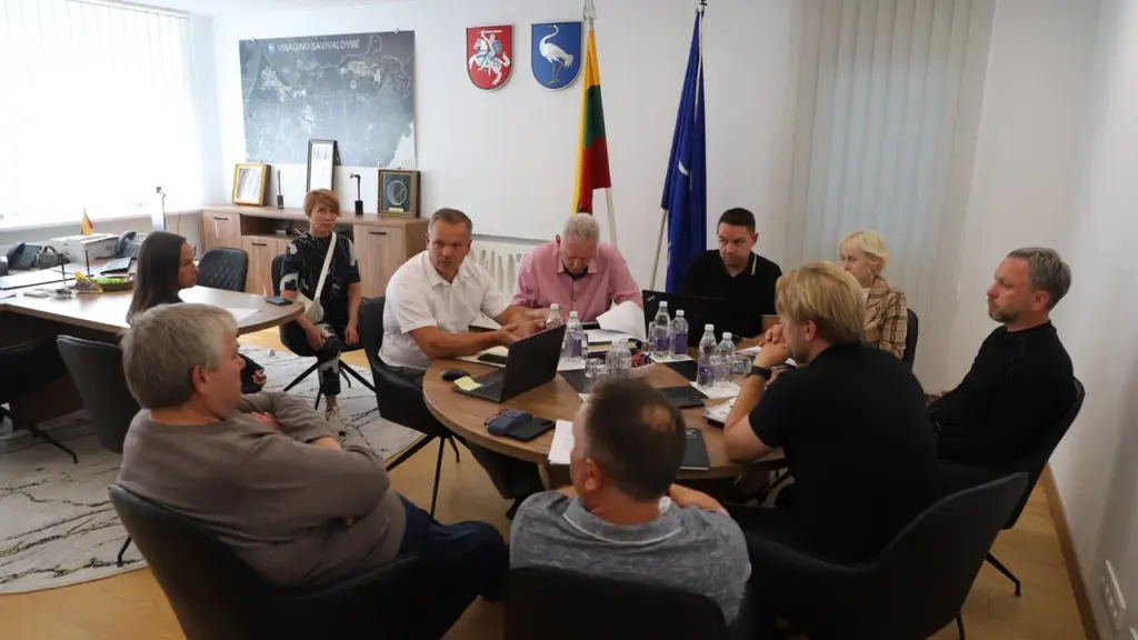 Grupė žmonių sėdi prie apvalaus stalo posėdžių salėje su Lietuvos ir savivaldybės vėliavomis.