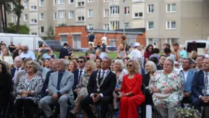 Grupė oficialių asmenų ir miestiečių sėdi lauke stebėdami renginį daugiabučių namų fone.