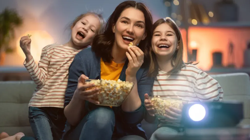 Šeima su vaikais smagiai žiūri filmą ir valgo spragėsius namų kino seanso metu.