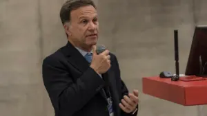 Vidutinio amžiaus vyras su kostiumu laiko mikrofoną ir kalba konferencijoje, stovėdamas prieš auditoriją.