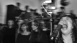 Moteris energingai dainuoja į mikrofoną šalia choro, fone matomas neryškus, judesio efektą turintis vaizdas.