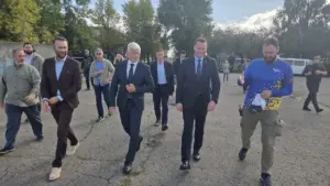 Grupė vyrų su kostiumais ir laisvalaikio apranga eina asfaltuota aikštele saulėtą dieną.